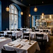 Innenansicht eines leeren, eleganten europäischen Restaurants am frühen Abend. Dunkelblaue Wände kontrastieren mit weiß gedeckten Tischen, Weingläsern und goldenen Pendelleuchten. Im Hintergrund ist eine glänzende Edelstahl-Passküche sichtbar. Große Rundbogenfenster lassen sanftes Tageslicht herein.