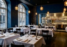 Innenansicht eines leeren, eleganten europäischen Restaurants am frühen Abend. Dunkelblaue Wände kontrastieren mit weiß gedeckten Tischen, Weingläsern und goldenen Pendelleuchten. Im Hintergrund ist eine glänzende Edelstahl-Passküche sichtbar. Große Rundbogenfenster lassen sanftes Tageslicht herein.