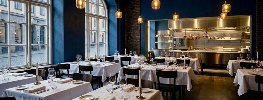 Innenansicht eines leeren, eleganten europäischen Restaurants am frühen Abend. Dunkelblaue Wände kontrastieren mit weiß gedeckten Tischen, Weingläsern und goldenen Pendelleuchten. Im Hintergrund ist eine glänzende Edelstahl-Passküche sichtbar. Große Rundbogenfenster lassen sanftes Tageslicht herein.