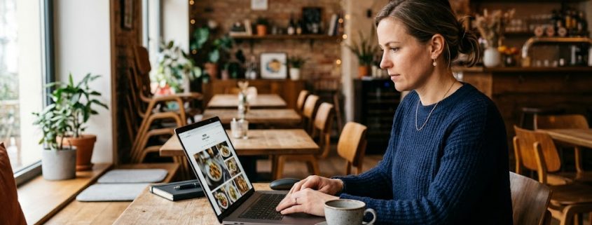 Eine Frau sitzt konzentriert in einem gemütlichen, leeren Restaurant an einem Holztisch und arbeitet an einem offenen Laptop. Neben dem Laptop steht eine Tasse Kaffee.