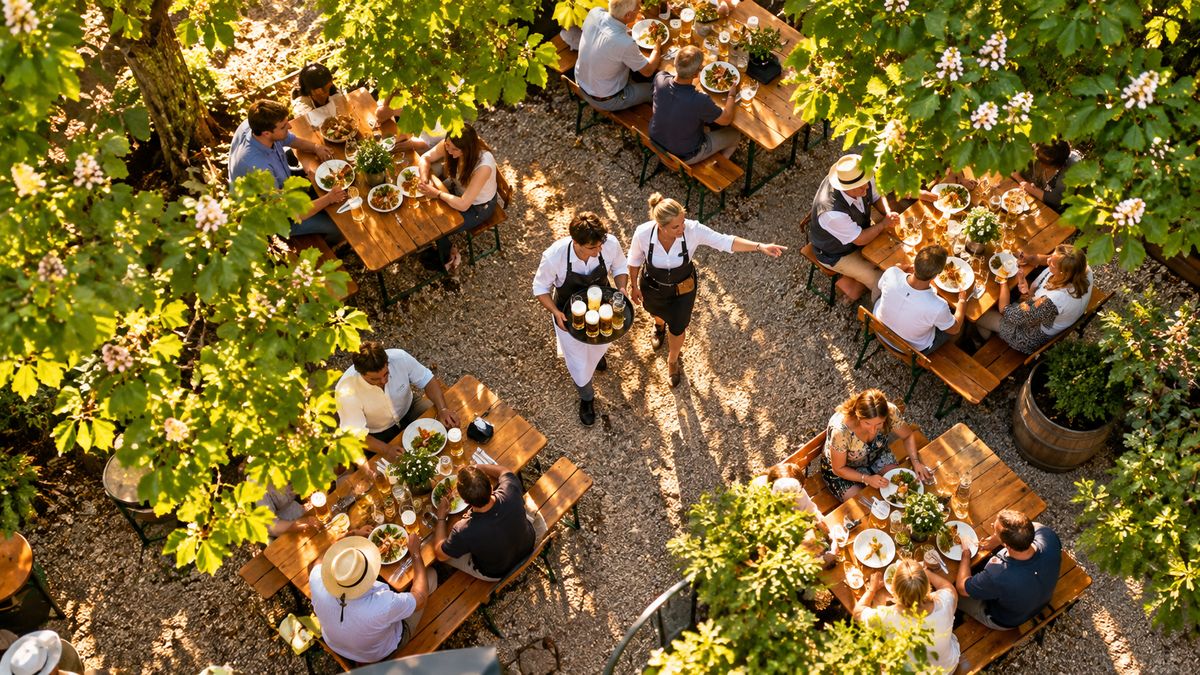Biergarten von oben: eine erfahrene Servicekraft zeigt einem Auszubildenden mit Bierkrügen auf dem Tablett den Weg zwischen den Tischen